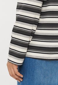 Svītrota garām piedurknēm krekls ar maiņām melnās un baltās svītrās; kakla izgriezums un piedurkņu galus ir ar gludu, noapaļotu apdari, kas kombinējas ar ziliem džinsiem.