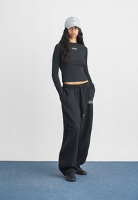 Haut à manches longues noir côtelé avec logo, associé à un pantalon de jogging noir ample arborant un logo blanc. Une casquette grise et des baskets noires complètent le look.