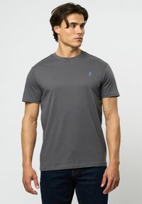 Grå bomulls t-skjorte med korte ermer, ribbet rund hals og blått logo på venstre bryst. Kombinert med mørke jeans.