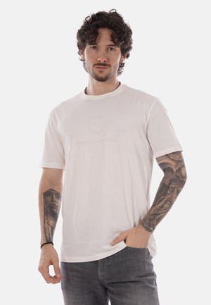Hombre con cabello rizado y brazos tatuados, vistiendo una camiseta blanca y vaqueros grises, posando con una mano en el bolsillo sobre un fondo blanco.