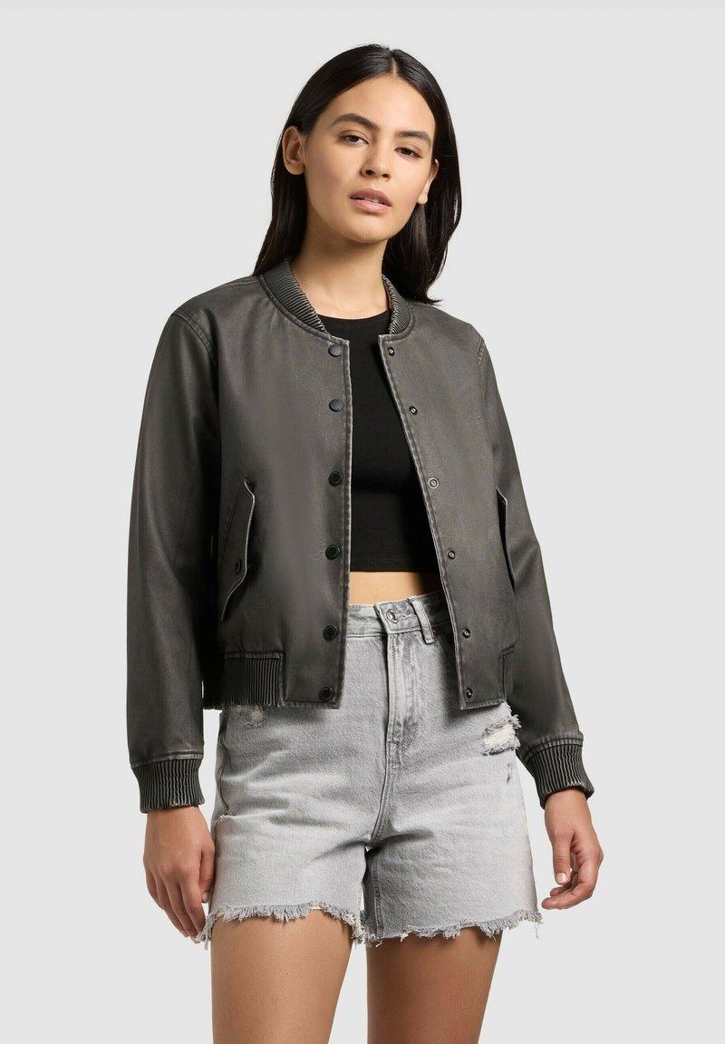 Femme portant une veste bomber gris foncé sur un crop top noir et un short en denim gris clair déchiré avec des bords effilochés.