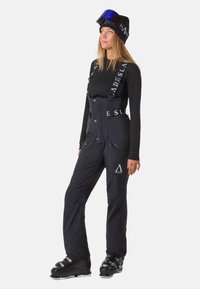 Zwarte skioverall met verstelbare banden, ritszakken en logo-accenten. Draagt een bijpassend zwart langemouwtop en skischoenen.