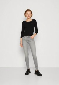 Junge Frau in einem schwarzen Langarmtop, grauen Skinny-Jeans und schwarzen Schuhen, die vor einem schlichten hellen Hintergrund steht, mit einer Hand in der Hosentasche.