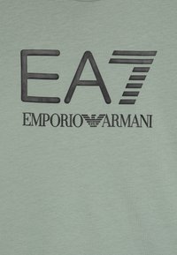 T-shirt vert clair avec un logo noir en gras "EA7 EMPORIO ARMANI", avec des détails distincts et une texture lisse. Design à manches courtes.
