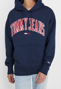 Felpa blu navy in cotone, con tasca a marsupio frontale, cordini e logo ricamato rosso "TOMMY JEANS". Indossati jeans azzurri.