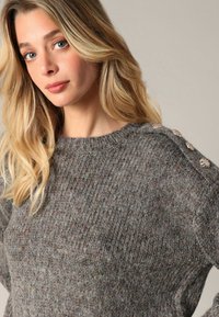 Pull en tricot gris avec une texture côtelée, encolure ronde et boutons décoratifs à l'épaule pour le détail.