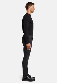 Chemise noire à manches longues, jean gris foncé ajusté et chaussures noires. La vue latérale montre un design minimaliste avec une texture lisse et des lignes épurées.