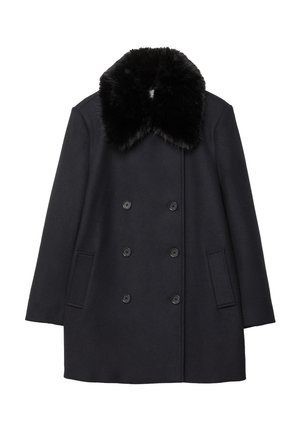 Personne portant un manteau noir croisé avec col en fourrure, un haut noir orné de dentelle, un jean large foncé et des chaussures noires, debout à l'intérieur.