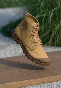 Zapatilla alta de lona amarilla con suela de goma marrón, cierre de cordones en la parte delantera y parche de logo en el lateral. Parte superior texturizada y materiales duraderos.