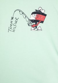 Světle zelené tričko s kreslenou postavičkou s rybářským prutem a textem "TOMMY HILFIGER" v černé barvě, umístěným vedle postavičky.