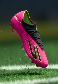 Φωτεινές ροζ και μαύρες ποδοσφαιρικές πατούσες με κίτρινο-πράσινες λεπτομέρειες που ανασηκώνουν γρασίδι σε έναν πράσινο αγωνιστικό χώρο κοντά σε μια λευκή γραμμή.