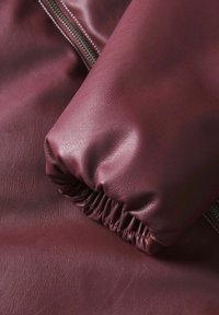 Blat burgundowej skóry, zbliżenie, z mankietem rękawa w fałdy i detalem zamka, gładka tekstura z delikatnym połyskiem.