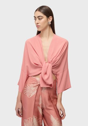 Kobieta o długich ciemnych włosach, nosząca duże srebrne kolczyki, luźną różową bluzkę związaną z przodu oraz szerokie spodnie w różowe kwiaty.