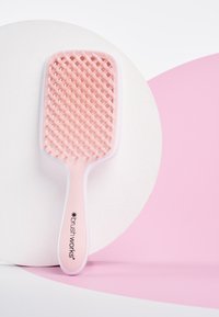 Rektangulär hårborste med ett strukturerat rosa gummihuvud, vit plastkant och ergonomiskt handtag. Har en logotyp på handtaget.