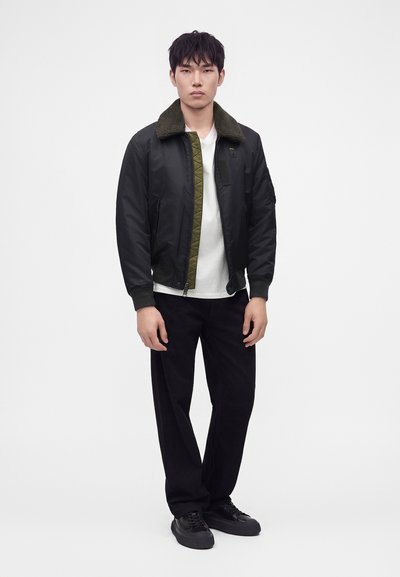 Homme portant une veste bomber noire avec une doublure verte sur une chemise blanche, pantalon noir et chaussures noires, debout devant un fond blanc uni.
