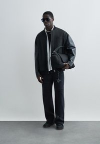 Fekete bombázó dzseki bőr ujjakkal, fehér inggel alatta, fekete farmernadrág szegecses díszítéssel, és fekete tornacipővel.