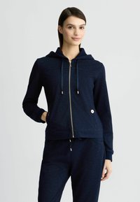 Felpa con zip blu navy realizzata in tessuto testurizzato, con cappuccio, zip dorata e lacci. Presenta un logo ricamato e tasche laterali.