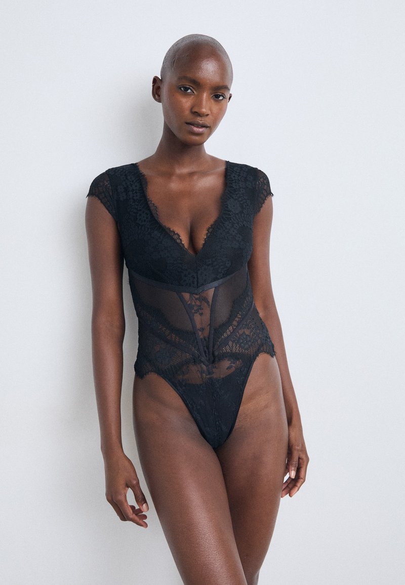 Black lace bodysuit with cap sleeves, deep V-neck, sheer panels, and a high-cut leg. Textured floral pattern throughout.
