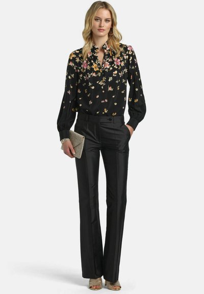 Femme portant un chemisier noir à fleurs avec des manches bouffantes, un pantalon noir coupé sur mesure, tenant une pochette dorée scintillante et des talons ouverts dorés.