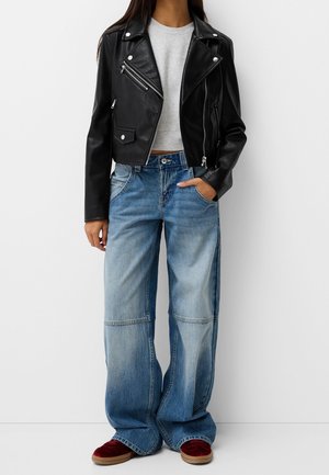 Personne portant une veste en cuir noire, un top court côtelé gris, un jean large bleu délavé et des baskets rouges, debout devant un fond blanc.
