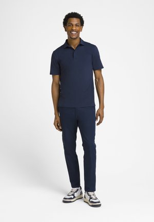 Smilende mand stående iført en navy blå polo shirt, matchende bukser og hvide sneakers med sorte accenter på en ensfarvet hvid baggrund.
