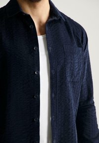 Man in een niet-knopopgemaakt marineblauwe corduroy shirt over een witte geribde tanktop, waarbij de textuur van de stof en de borstzak zichtbaar zijn.