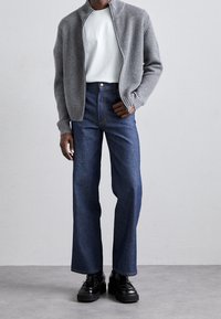 Grå ribbad zip-up cardigan över en vit T-shirt, tillsammans med högmidjade blå jeans. Svarta skor med en chunky design.