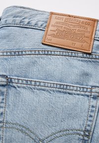 Ljusblå denimjeans har en läderpatch med präglad text. Tyget visar en strukturerad väv och kontrasterande gul söm.