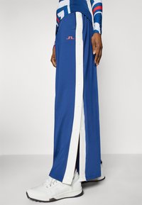 Modrá dlhá sukňa so zapínaním na bočných cvokoch, bielymi akcentmi a logom. Vhodne kombinovaná s bielymi športovými topánkami pre športový vzhľad.