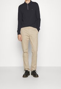 Uomo che indossa un polo nero a maniche lunghe, chinos beige e scarpe nere stringate, in piedi davanti a uno sfondo bianco.