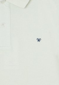 Polo verde chiaro, tessuto testurizzato, tre bottoni al collo, con un piccolo logo delle forbici blu navy sul lato sinistro del petto.