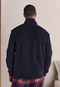 Marineblaue Fleecejacke mit Stehkragen, langen Ärmeln und strukturierter Stoffstruktur. Wird über karierten Pyjamahosen getragen, Ansicht von hinten.