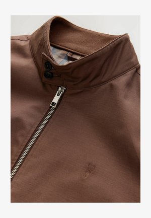 Braune Jacke mit hohem Kragen, Reißverschluss und zwei schwarzen Knöpfen. Glattes Gewebe mit dezenter Textur und gesticktem Logo-Detail.