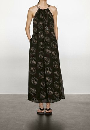 Kobieta ubrana w czarną, bezrękawnikową suknię maxi z dużymi okrągłymi wzorami i czarne sandały, stojąca z rękami w kieszeniach.