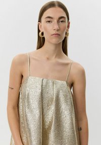 Metallisk guldklänning med spaghetti-band, som har en texturerad yta och en lös, flödande silhuett. Djärva guldörhängen i form av ringar kompletterar outfiten.