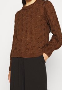 Brązowy sweter dziergany z teksturowanym wzorem w wypukłe diamenty, z prążkowanym dekoltem i mankietami oraz luźnym krojem.