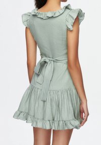 Hellgrünes Leinenkleid mit Rüschensäumen, tailliertem Oberteil und schwingendem Rock. Das Bindeband im Rücken verleiht dem Design Struktur.