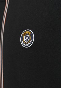 Tissu noir avec des rayures verticales rouges, blanches et bleues et un patch logo rond "Premium Jack & Jones" en blanc, orange et bleu marine.