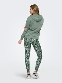Femme de dos portant un sweat à capuche vert, des leggings imprimé léopard vert et noir, et des baskets blanches, debout devant un fond uni.