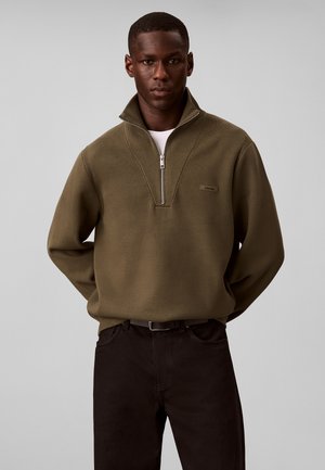 Ung mand iført en olivengrøn sweater med kvart lynlås over en hvid skjorte og mørke bukser mod en ensfarvet grå baggrund.