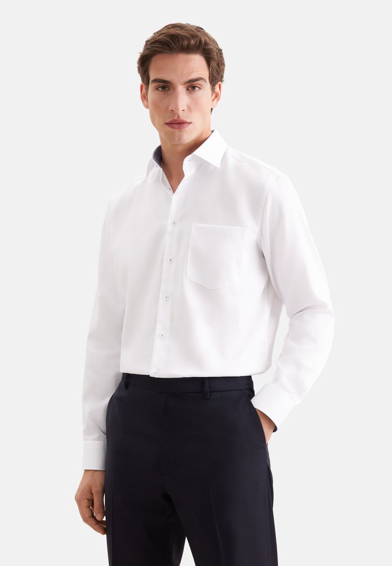 Homme portant une chemise blanche à manches longues avec une poche devant et un pantalon foncé, debout avec une main dans la poche.
