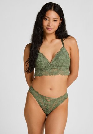 Vrouw met lang zwart haar, gekleed in een groene kanten bralette en bijpassende kanten slip, staand tegen een effen lichtgrijze achtergrond.