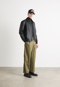 Veste en cuir noir par-dessus un haut noir, avec un col et une fermeture à boutons. Un pantalon ample vert olive et des chaussures noires complètent la tenue. Une casquette noire sur la tête.