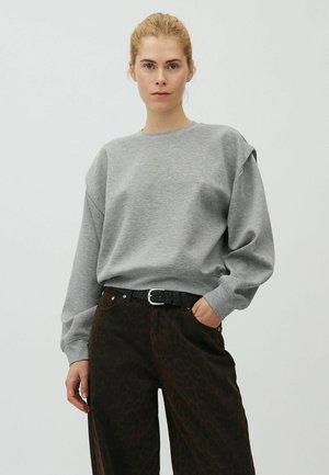 Kvinde med blondt hår iført en grå langærmet sweatshirt og mørke mønstrede bukser, stående med den ene hånd i lommen mod en ensfarvet baggrund.