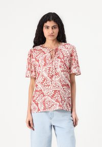 Kortermet bluse i rustfarge med hvit blomstermønster. Har en nøkkelhull-hals og mykt stoff. Kombinert med lyseblå, løstsittende jeans.