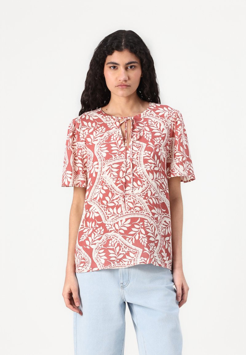 Kortermet bluse i rustfarge med hvit blomstermønster. Har en nøkkelhull-hals og mykt stoff. Kombinert med lyseblå, løstsittende jeans.