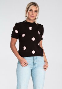 Kobieta w czarnej krótkiej bluzce z różowymi wzorami kwiatowymi i jasnoniebieskich dżinsach, stojąca na tle jasnego, jednolitego tła.