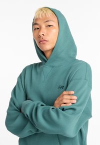 Sudadera con capucha turquesa de material texturizado y acanalado. Cuenta con capucha y pequeño logo en el pecho. Mangas largas, ajuste relajado y escote en forma de V.