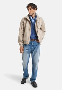 Beige Bomberjacke über einem blauen Reißverschluss-Sweatshirt, kombiniert mit blauen Jeans und dunklen Schuhen, dazu ein brauner Gürtel und strukturierter Stoff.