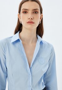 Camisa de botones azul claro con cuello, con un logo bordado en el pecho. Fabricada en tela suave con un corte clásico.